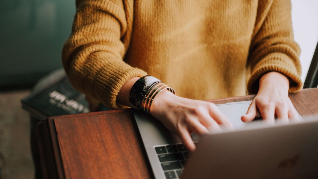person in orange sweater is typing on a laptop