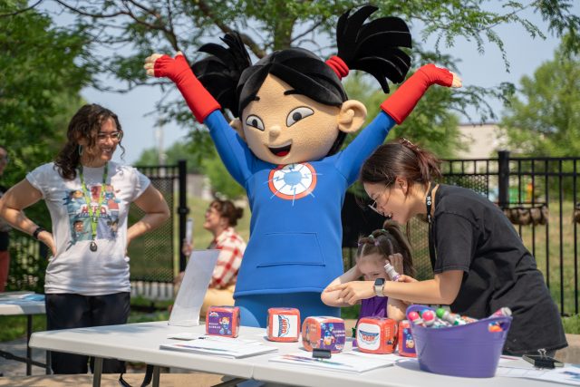 a costume character of Sara Snap from Hero Elementary stretches her arms out while people are engaging in arts and crafts